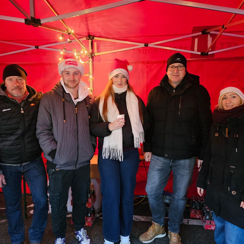 Fünf Personen, die warm gekleidet sind und Wintermützen tragen, stehen gemeinsam unter einem roten Zelt. Sie alle lächeln und scheinen für ein Foto zu posieren.