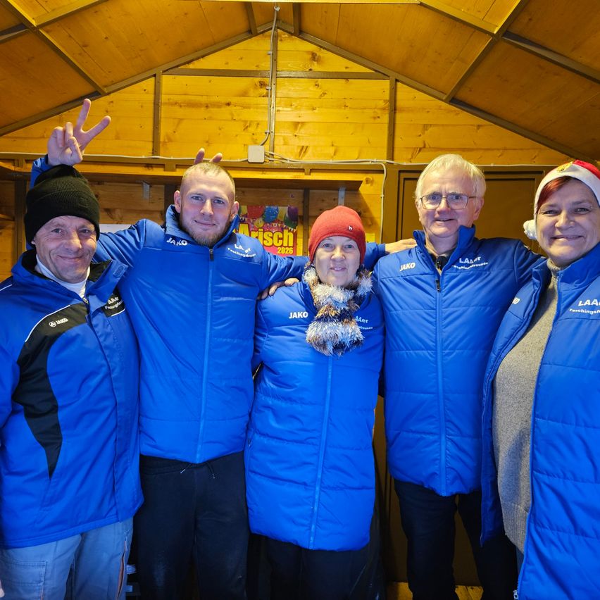 Fünf Menschen in blauen Jacken und Winterkleidung posieren für ein Foto in einer Holzstruktur. Sie haben Lächeln auf den Gesichtern. Hinter ihnen befindet sich eine Holzwand mit einer Steckdose.