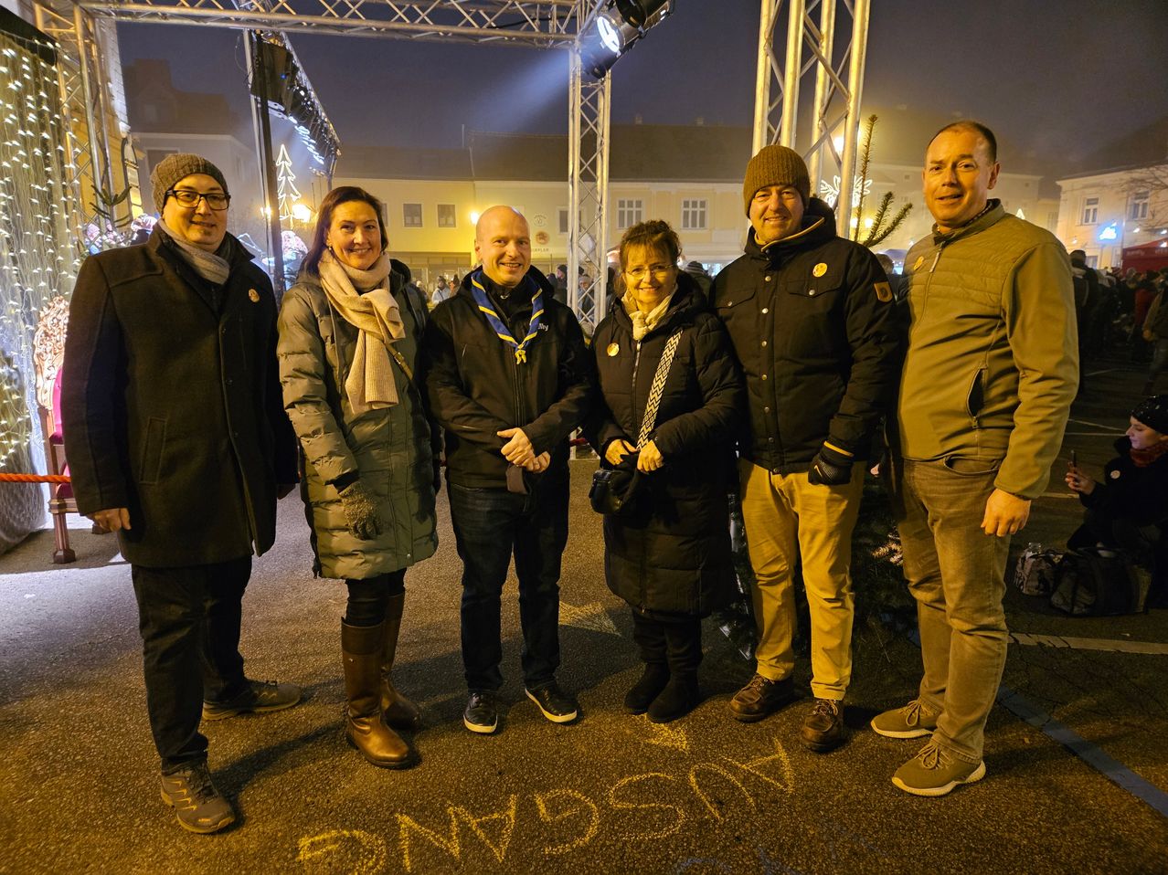 Eine Gruppe von Menschen in Winterkleidung posiert nachts vor einem Gebäude für ein Foto, auf dem Boden steht das Wort 'SMWSYN' geschrieben.