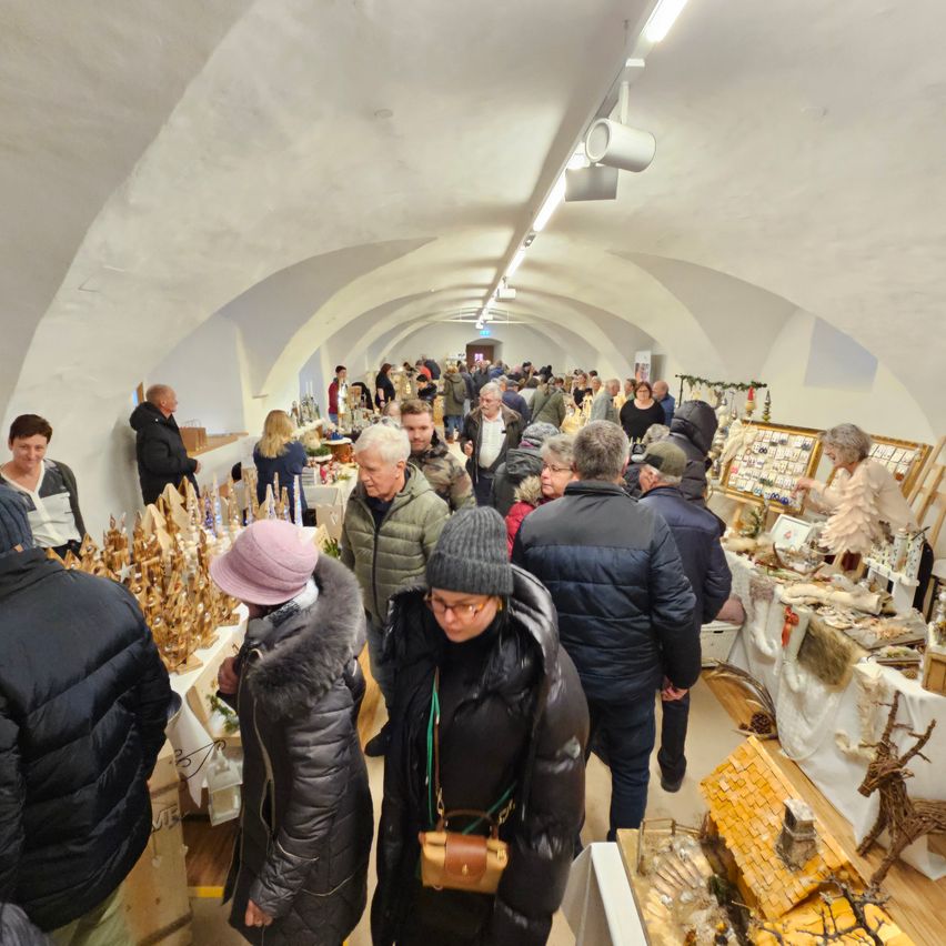 Ein Indoor-Markt mit gewölbten Decken, wo zahlreiche Menschen verschiedene Stände durchstöbern und Gegenstände wie Holzfiguren und Handwerkskunst präsentieren. Die Decke ist mit Lichtern versehen und die Wände sind mit verschiedenen Kunstwerken geschmückt.