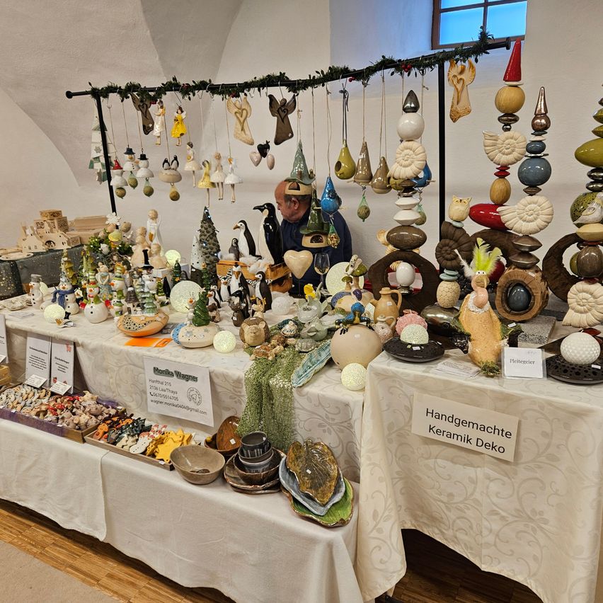 Ein Tisch voller handgefertigter Keramikdekorationen hängt von einer Stange. Ein Mann sitzt hinter dem Tisch, umgeben von verschiedenen Keramikartikeln. Ein Schild auf dem Tisch lautet 'Handgemacher Keramik Deko'.