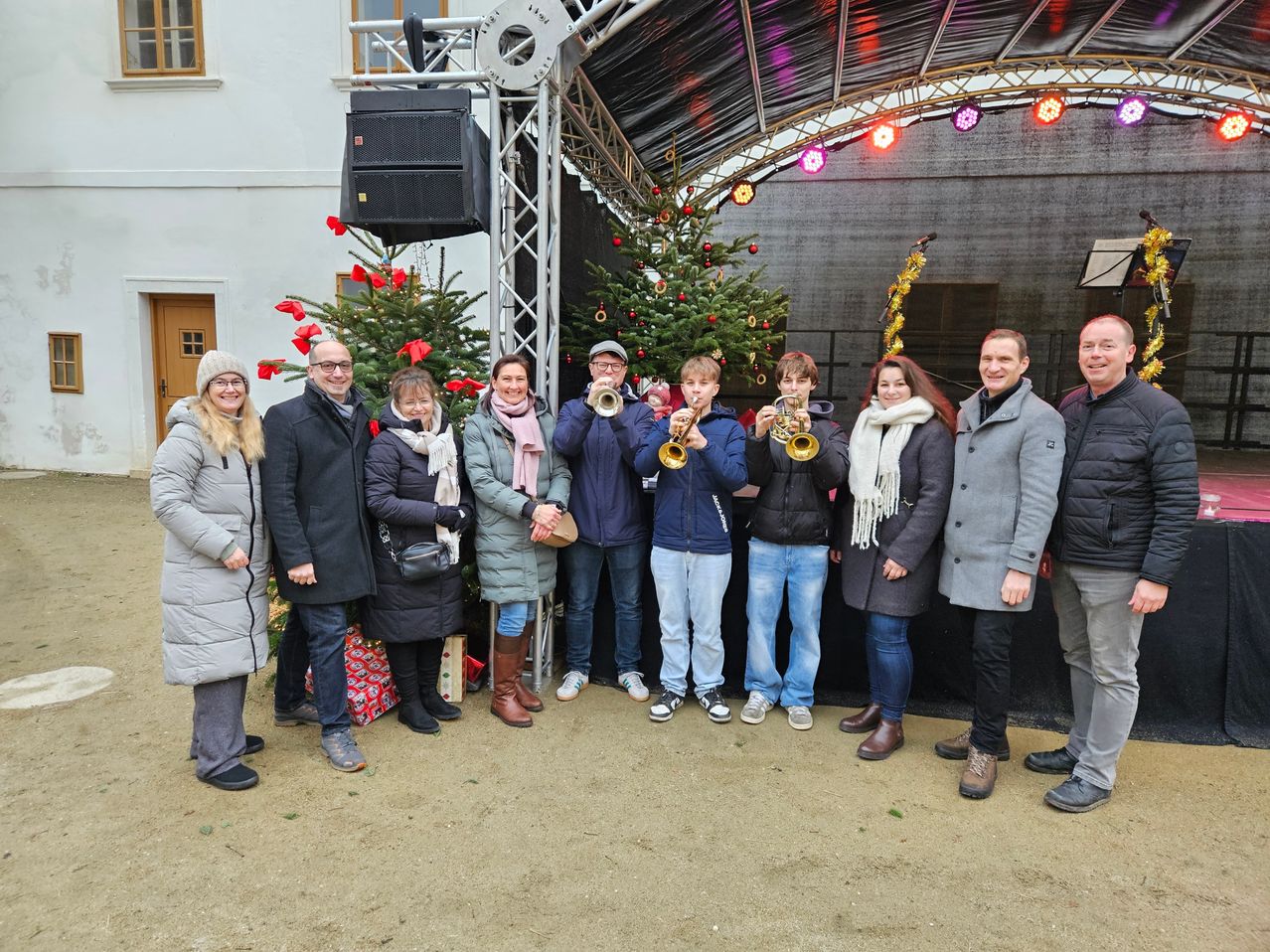 Eine Gruppe von Menschen steht vor einem geschmückten Weihnachtsbaum und hält Trompeten. Sie tragen Winterkleidung und lächeln für ein Foto.