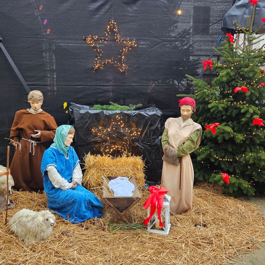 Eine Krippe mit einem Stern auf schwarzem Hintergrund. Ein Weihnachtsbaum mit roten Schleifen steht in der Nähe. Maria und Josef knien neben der Krippe mit einem Baby darin. Ein Schaf ist ebenfalls in der Nähe.