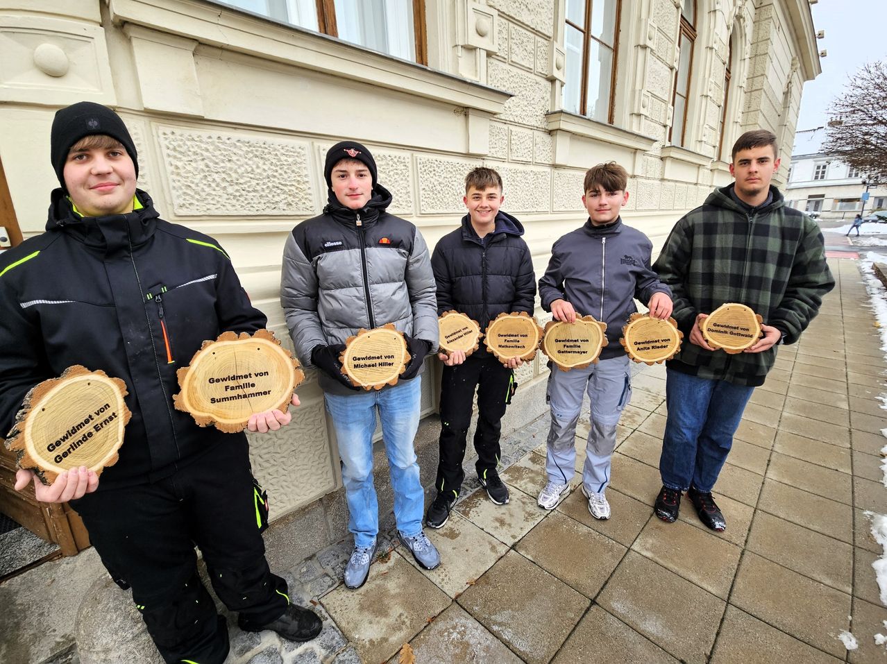 Fünf junge Männer stehen vor einem Gebäude und halten Holztafeln in den Händen. Jede Tafel hat eine einzigartige Botschaft, die darauf graviert ist. Sie sind alle warm gekleidet, was darauf hindeutet, dass es draußen kalt sein könnte.