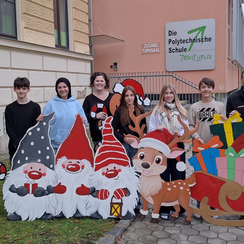 Eine Gruppe von Menschen posiert für ein Foto vor einem Gebäude mit dem Schild Die Polytechnische Schule. Sie halten weihnachtliche Ausschnitte, darunter Zwerge, ein Rentier und einen Schlitten.