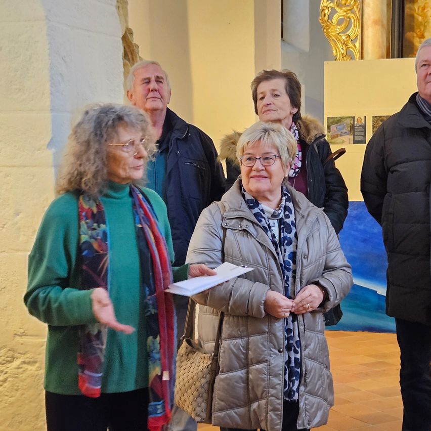 Eine Gruppe älterer Erwachsener steht in einem Gebäude in der Nähe einer Wand, zwei Frauen halten Papiere, während andere in die Kamera schauen. Ein blaues Gemälde und ein goldener Rahmen sind im Hintergrund zu sehen.