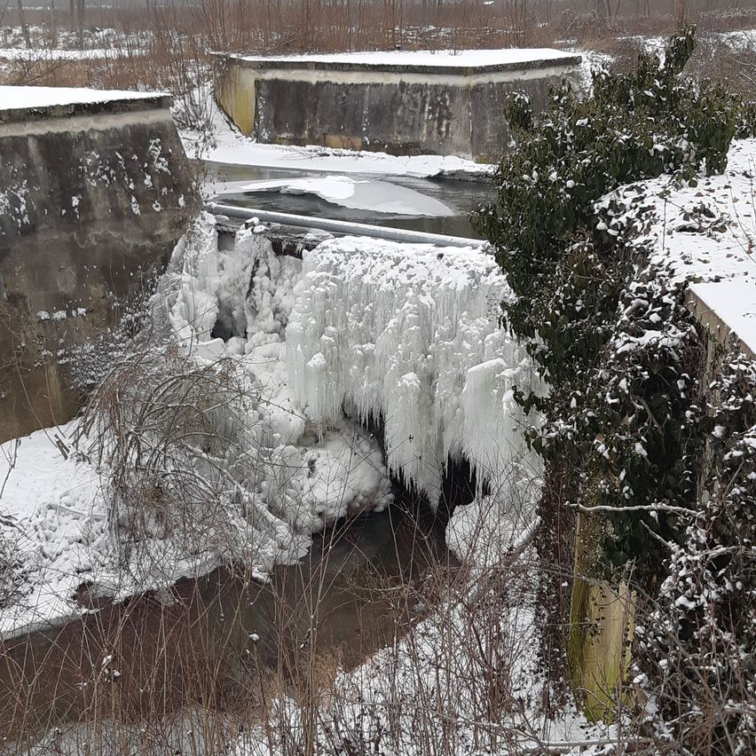 Schnee bedeckt einen Wasserfall mit eisigen Formationen, der durch einen Betonkanal in einer Winterlandschaft fließt.