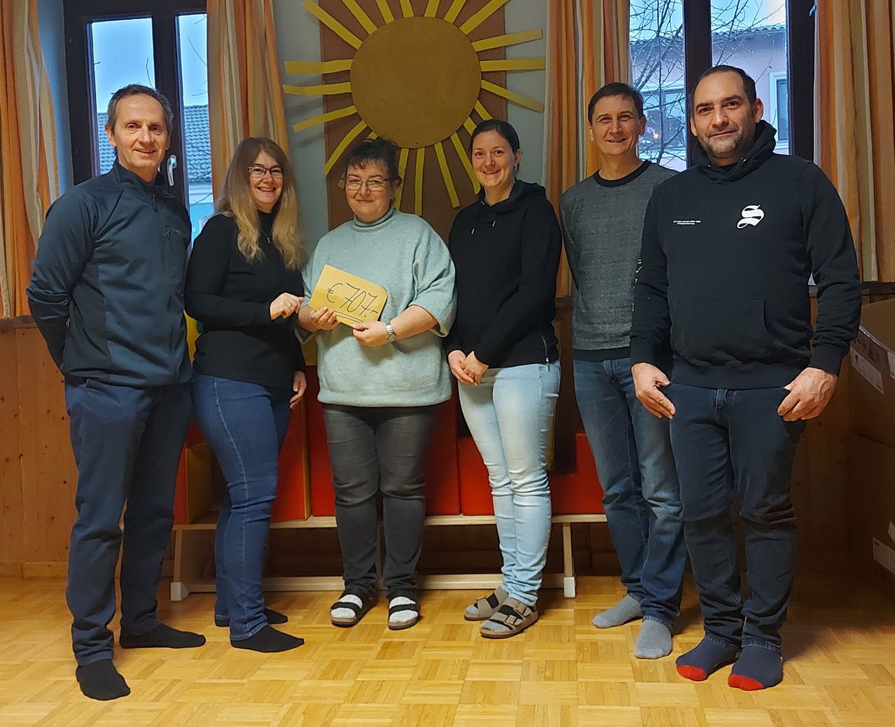 Eine Gruppe von fünf Erwachsenen, vier Frauen und einem Mann, stehen zusammen in einem Raum mit einem Holzboden. Die Frau in der Mitte hält ein Schild mit der Aufschrift '407 €'. Sie lächeln und scheinen für ein Foto zu posieren. Hinter ihnen befindet sich eine Wand mit einem Sonnendesign und einem Fenster.