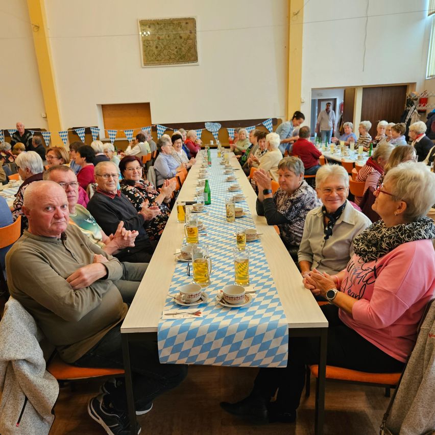 Eine Gruppe älterer Menschen sitzt an einem langen Tisch mit blau-weiß karierten Tischdecken. Sie haben Gläser Bier und Tassen auf Untertassen vor sich. Sie klatschen und lächeln.