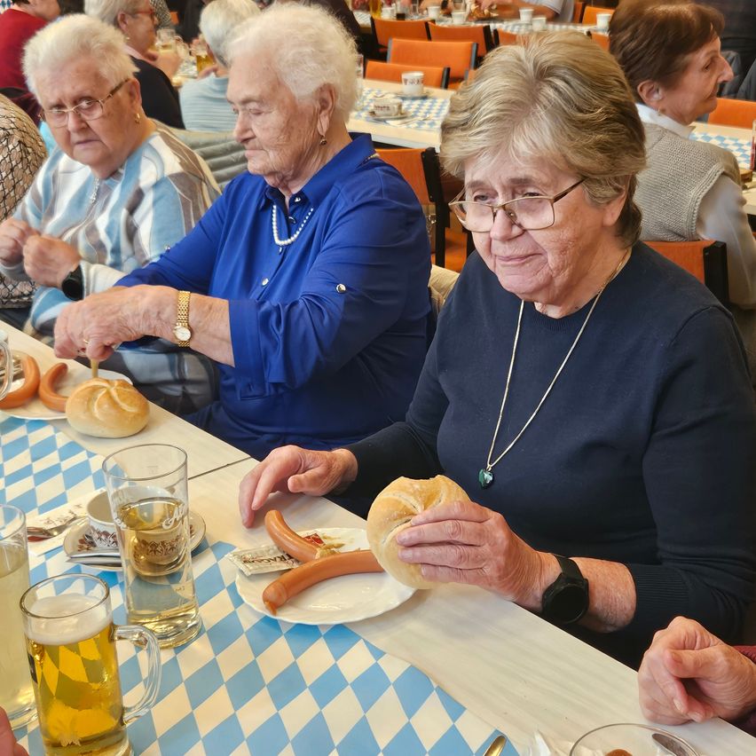 Einige ältere Frauen sitzen an einem Tisch und genießen Essen und Getränke. Eine Frau in einem blauen Hemd hält ein Brötchen und eine Wurst. Eine andere Frau in einem blauen Hemd und einer Uhr hält ein Brötchen.