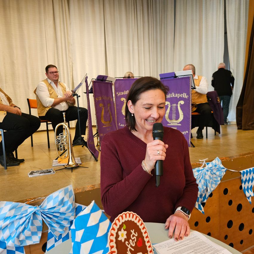 Eine Frau steht an einem Tisch mit einem Kuchen und spricht in ein Mikrofon. Hinter ihr spielen Musiker auf der Bühne, geschmückt mit traditionellen Dekorationen.