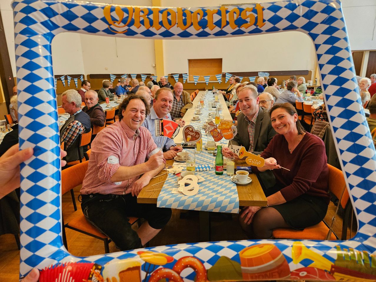Eine Gruppe von Menschen auf einem deutschen Oktoberfest, lächelnd und verschiedene Gegenstände haltend, sitzen um einen langen Tisch herum, der mit blau-weiß karierten Tischdecken dekoriert ist.