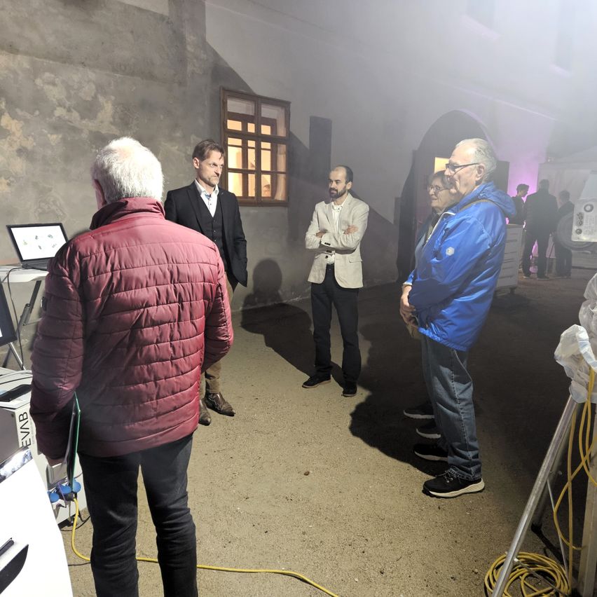 Fünf Männer stehen in einem schwach beleuchteten Raum. Ein Mann schaut auf einen Laptop, während andere die Arme verschränkt haben.