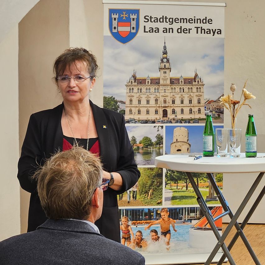 Eine Frau steht neben einem Plakat, das Laa an der Thaya bewirbt, mit einem Mann, der vor ihr sitzt, während ein Tisch mit Gläsern und Flaschen hinter ihnen steht.