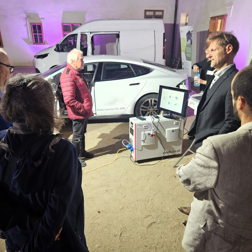 Eine Gruppe von Menschen steht um ein Tesla-Elektroauto herum. Ein Mann in einer roten Jacke spricht mit einem Mann im schwarzen Anzug. Ein Monitor steht auf einem Ständer.