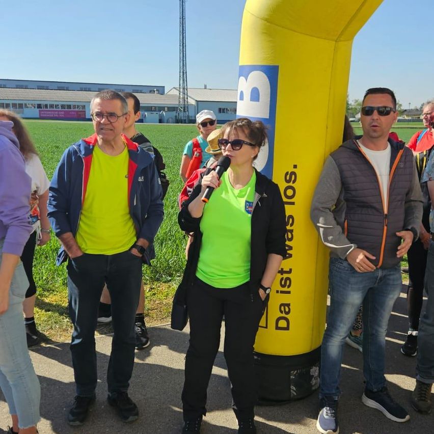 Eine Gruppe von Menschen steht in der Nähe eines gelben Bogens mit der Aufschrift 'Daist was los.' Eine Frau spricht in ein Mikrofon. Im Hintergrund befinden sich Gebäude und ein Feld.