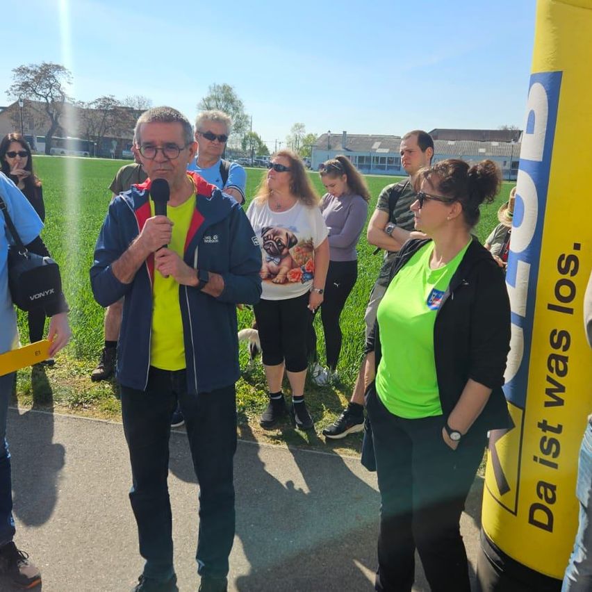 Eine Gruppe von Menschen versammelt sich auf einem Feld. Ein Mann spricht in ein Mikrofon, während eine Frau neben ihm steht. Andere Personen stehen herum und beobachten. Im Hintergrund sind Gebäude und Bäume zu sehen.