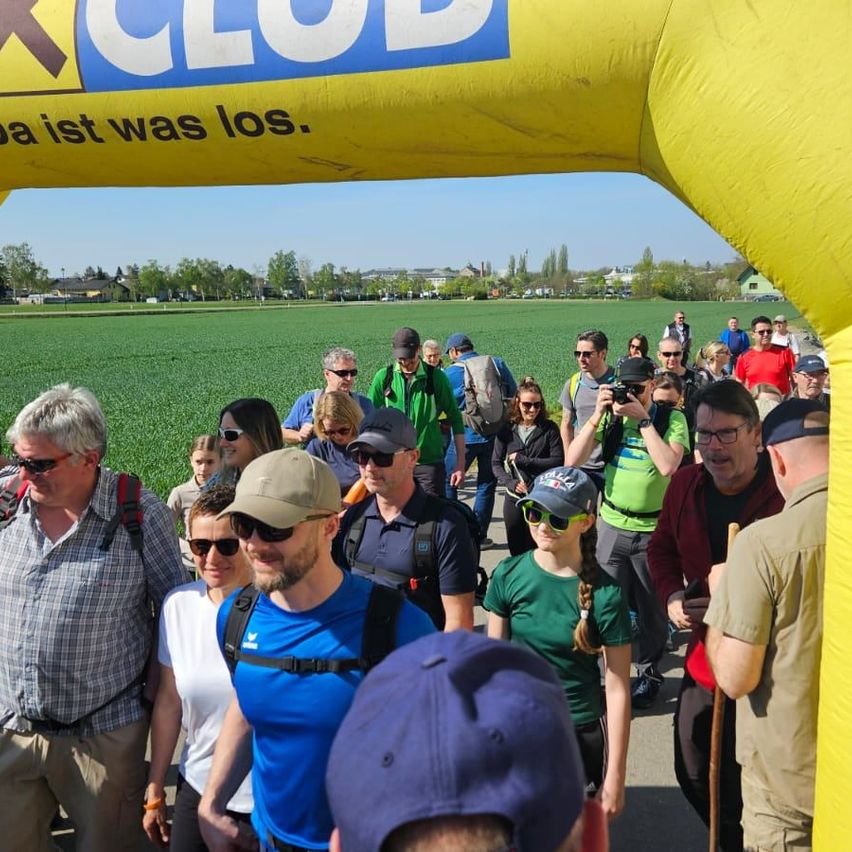 Eine Gruppe von Menschen in Outdoor-Ausrüstung geht durch einen gelben Bogen mit X Club darauf. Sie sind auf einem Feld mit Bäumen und Gebäuden im Hintergrund.