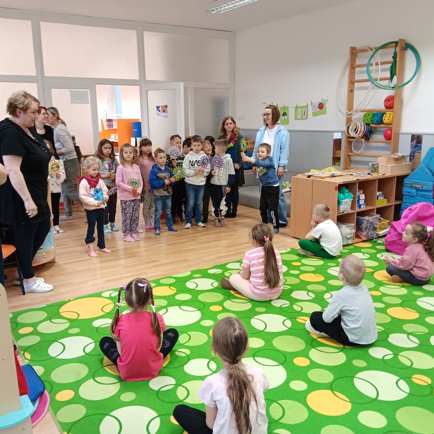 Eine Gruppe von Kindern sitzt auf dem Boden mit einem grünen Teppich. Sie halten Blumen. Zwei Frauen stehen neben ihnen. Es gibt eine Holzleiter mit Spielzeug auf der Seite.