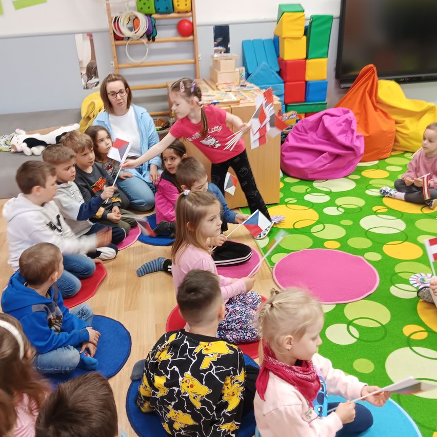 In einem Klassenzimmer sitzt eine Gruppe von Kindern in einem Kreis um eine Frau, die Flaggen hält. Einige von ihnen halten Flaggen und schauen die Frau an. Der Raum hat bunte Beanbags, Spielzeug und einen großen Bildschirm.