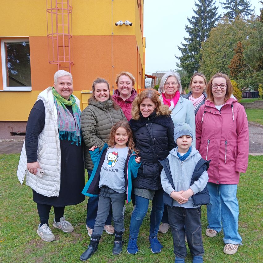 Eine Gruppe von Frauen und Kindern steht auf einem Rasen vor einem gelben Gebäude. Bäume und Pflanzen sind im Hintergrund.