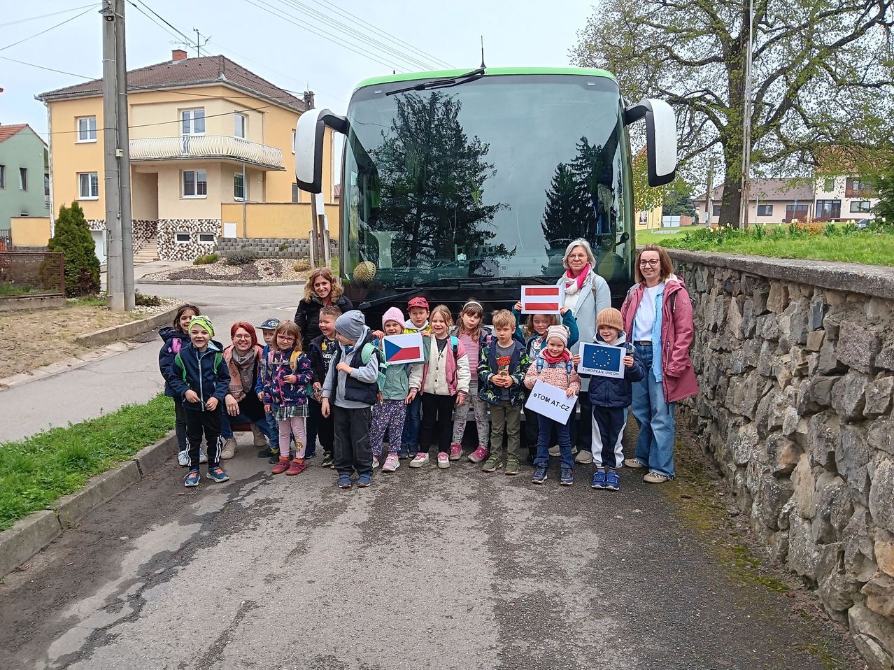 Eine Gruppe von Kindern und zwei Frauen steht vor einem Bus mit Flaggen. Die Kinder tragen Jacken und Turnschuhe. Die Frauen halten Fahnen mit den Texten 'Europäische Union' und 'TOM AT CZ'. Dahinter steht ein Haus mit Balkon.