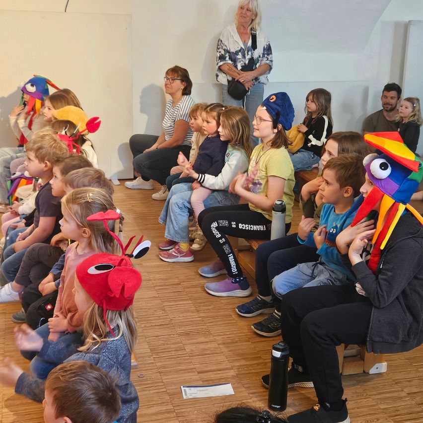 Kinder und Erwachsene sitzen in einem Raum. Kinder tragen Tiermützen. Eine Erwachsene steht und hält eine Tasche. Ein Kind trägt eine blaue Mütze und eine Halskette.