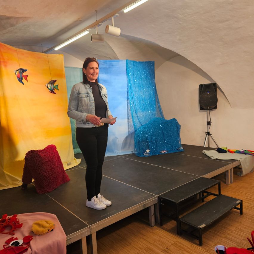 Eine Frau steht auf einer Bühne und hält ein Papier in der Hand, umgeben von farbenfrohen Kulissen und Requisiten. An der Wand ist ein Lautsprecher angebracht.