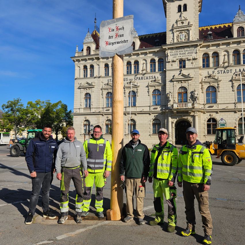Fünf Männer in Warnschutzkleidung posieren vor einem Gebäude mit einem Schild, das 'Ein Hoch der Franzl Bürgermeister' besagt.