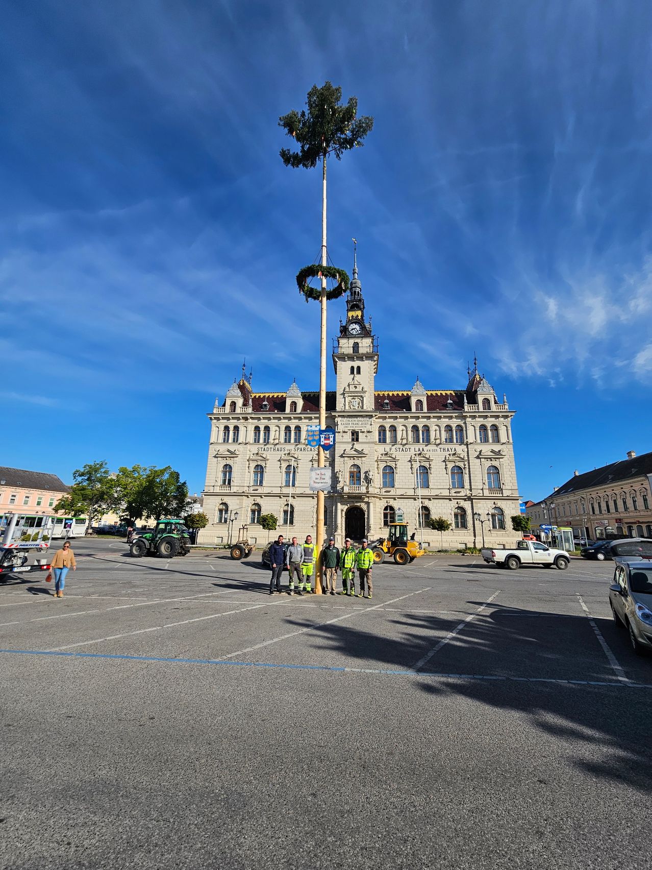 Eine Gruppe von Menschen versammelt sich vor einem großen Gebäude mit einem Glockenturm und einem hohen Pfahl mit einem Kranz, unter einem klaren blauen Himmel.