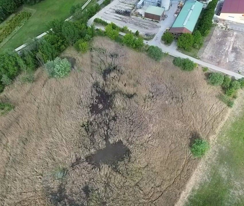 Luftaufnahme eines grasigen Feldes mit etwas Grün, einem unbefestigten Weg und mehreren Gebäuden und Strukturen im Hintergrund.