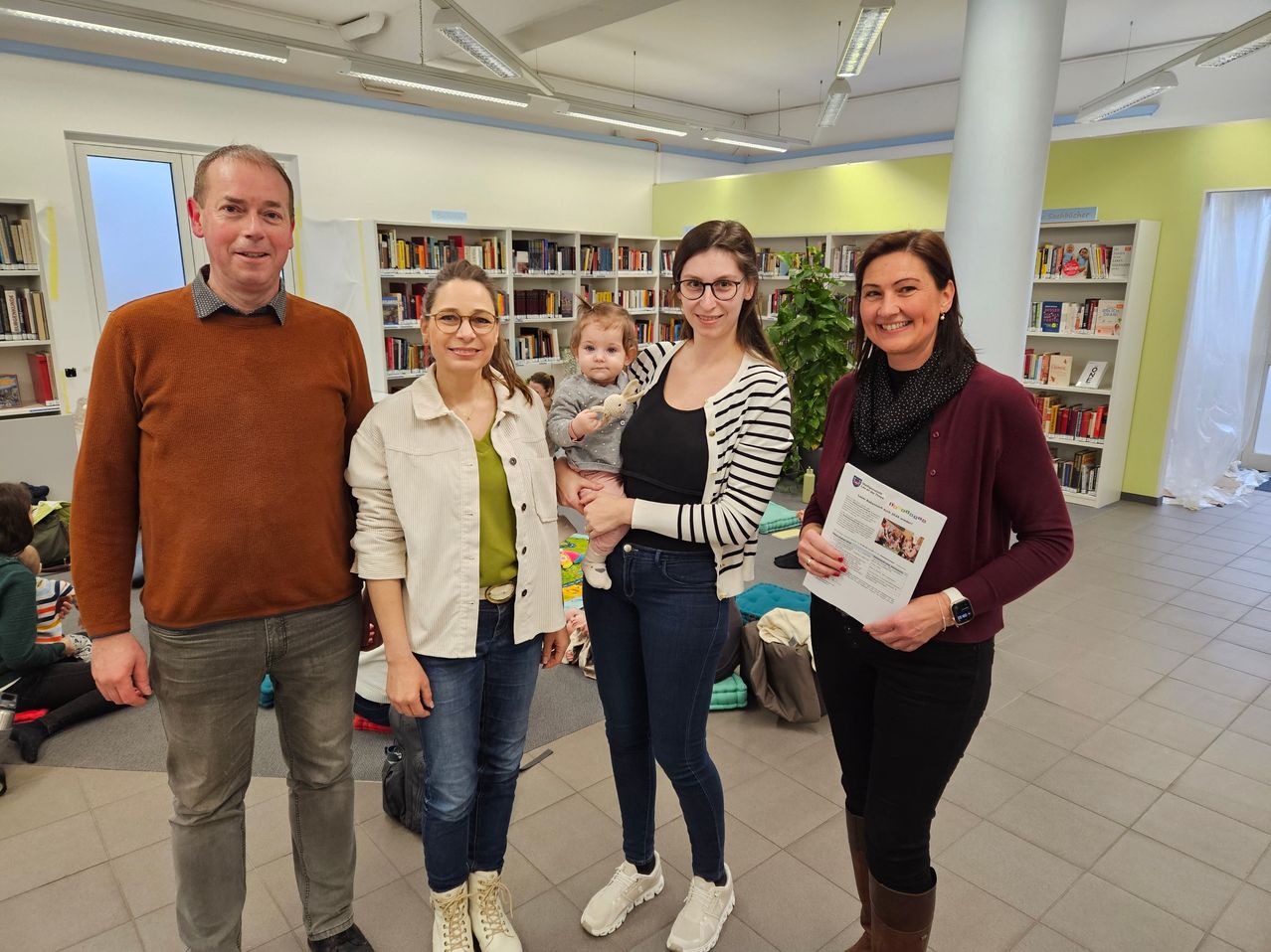 Eine Gruppe Erwachsener und ein Baby in einer Bibliothek. Sie lächeln und posieren für ein Foto. Hinter ihnen stehen Regale voller Bücher.
