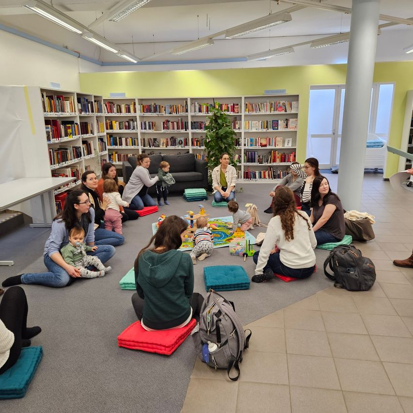 In einer Bibliothek sitzen mehrere Frauen und Kinder auf Kissen auf dem Boden. Eine Frau liest den Kindern vor. Im Hintergrund befinden sich Bücherregale.