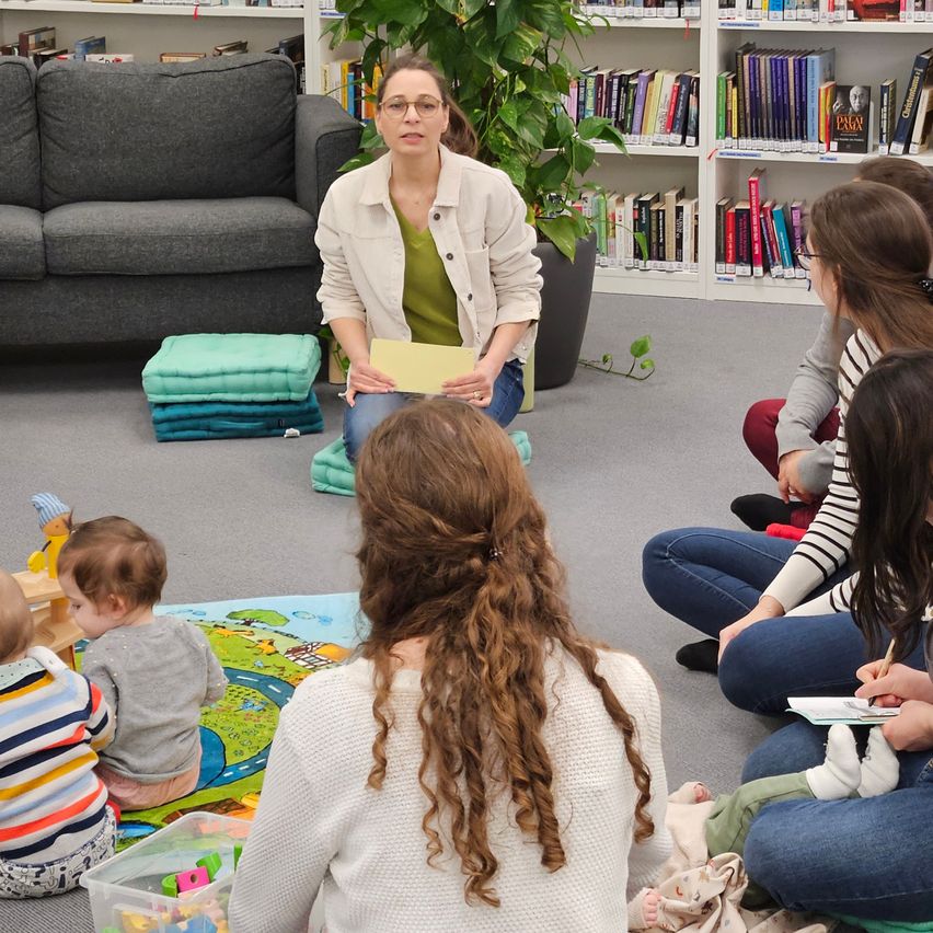 Eine Frau liest von einem gelben Papier vor einer Gruppe von Erwachsenen und Kindern, mit einer großen Couch, Büchern und einem Topfpflanzen im Hintergrund.