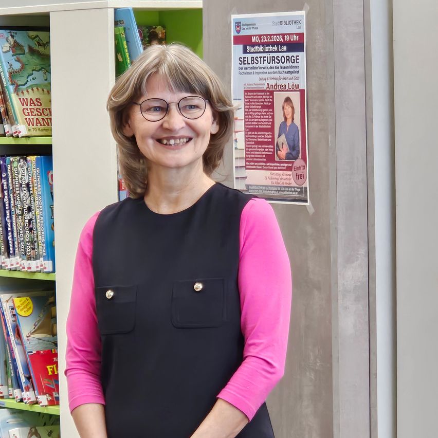 Eine Frau mit Brille und einem schwarzen und rosa Oberteil steht vor einem Bücherregal und einem Plakat.