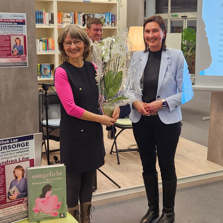 Zwei Frauen stehen in einer Bibliothek, eine hält einen Blumenstrauß und die andere lächelt. Ein Buch mit dem Titel 'Sattgeliebt' steht auf einem Tisch, mit Plakaten an der Wand. Ein Mann sitzt hinter ihnen auf einem Stuhl.