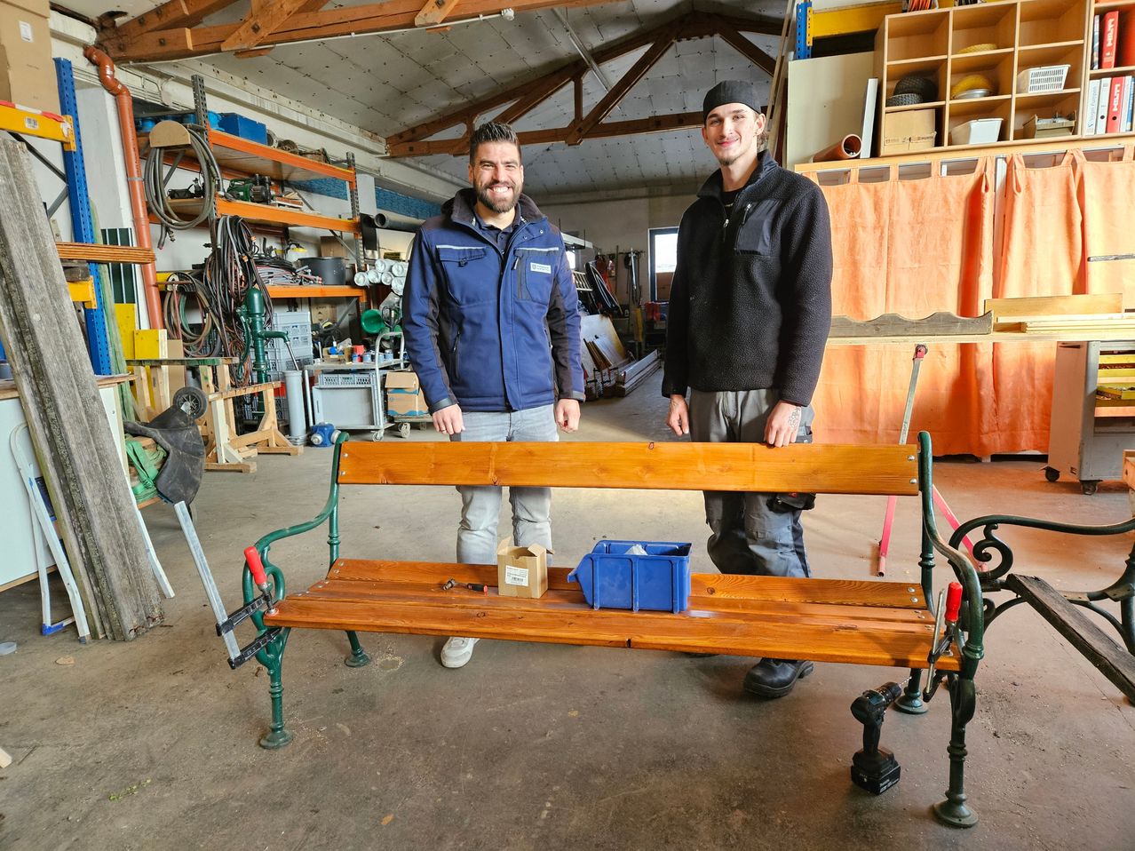 Zwei Männer stehen neben einer Holzbank in einer Werkstatt, einer trägt eine blaue Jacke und der andere eine schwarze Jacke. Sie sind von verschiedenen Werkzeugen und Materialien umgeben.