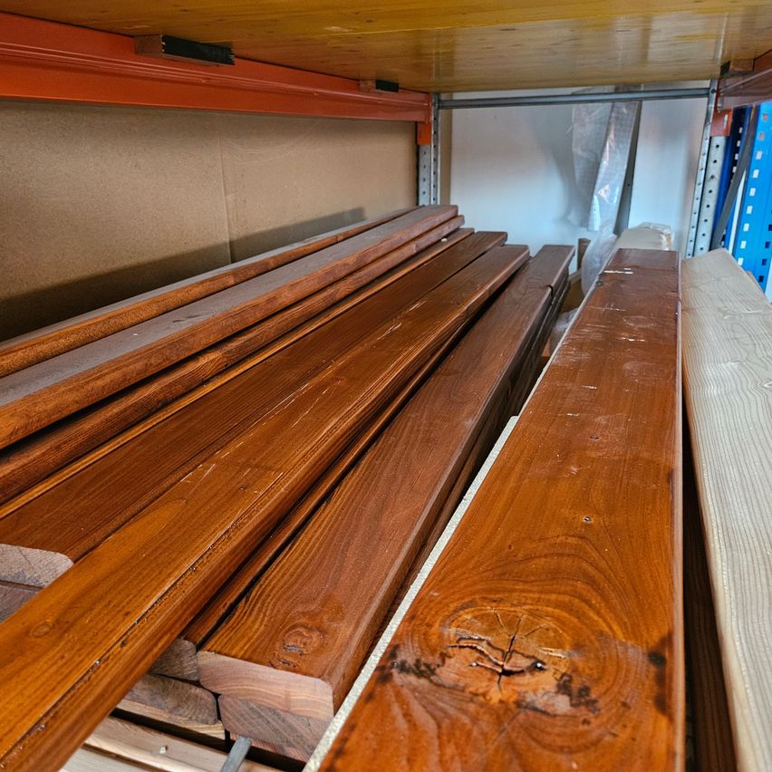 Eine Nahaufnahme verschiedener Holzbretter, die auf einem Lagerregal gestapelt sind. Die Bretter zeigen verschiedene Brauntöne und sind ordentlich angeordnet.