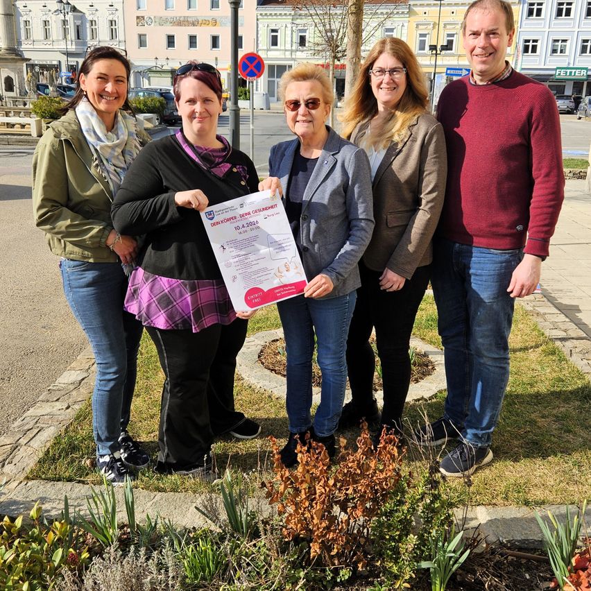 Fünf Erwachsene stehen lächelnd in einem Park. Eine Frau hält ein Schild mit Text und Logo. Sie sind von Grünflächen und Gebäuden umgeben.