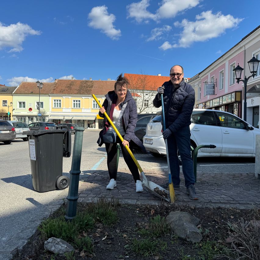 Zwei Personen reinigen eine Straßenecke. Sie haben Besen und lächeln. Ein Mülleimer steht in der Nähe. Im Hintergrund befinden sich Gebäude und geparkte Autos.