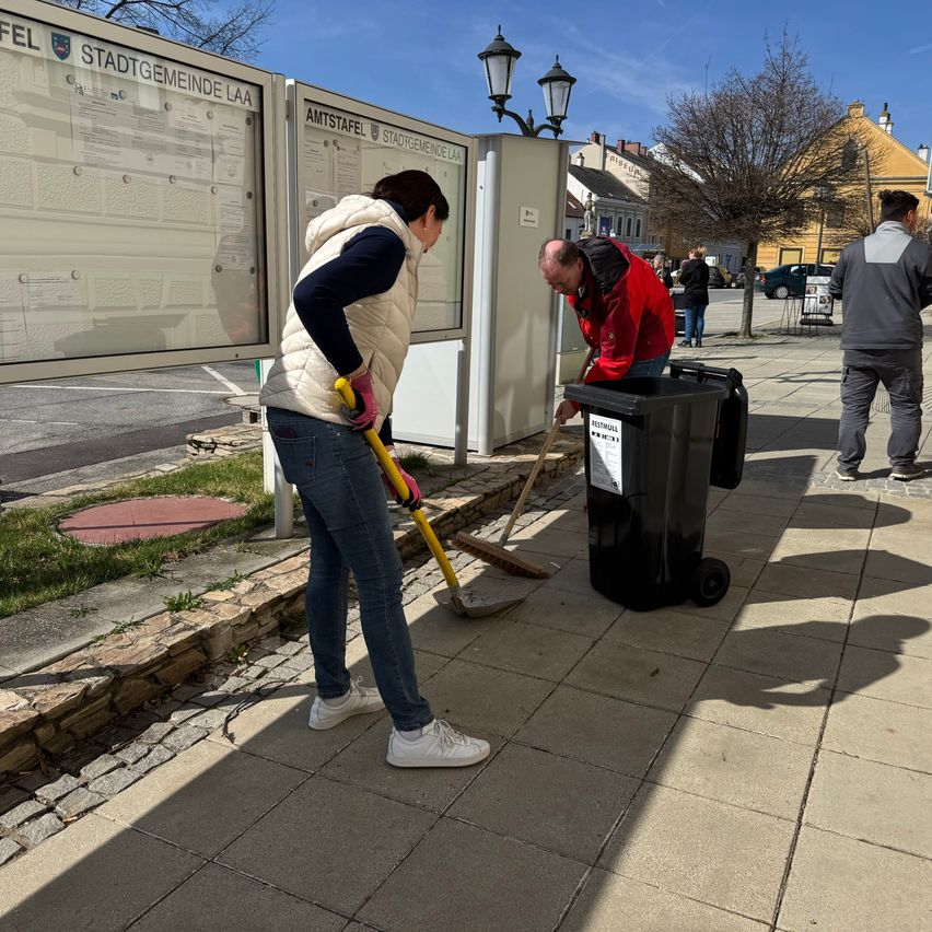 Zwei Personen reinigen einen Bürgersteig mit Besen. Ein Mülleimer ist in der Nähe. Eine weitere Person steht. Der Hintergrund umfasst Gebäude und einen Baum.