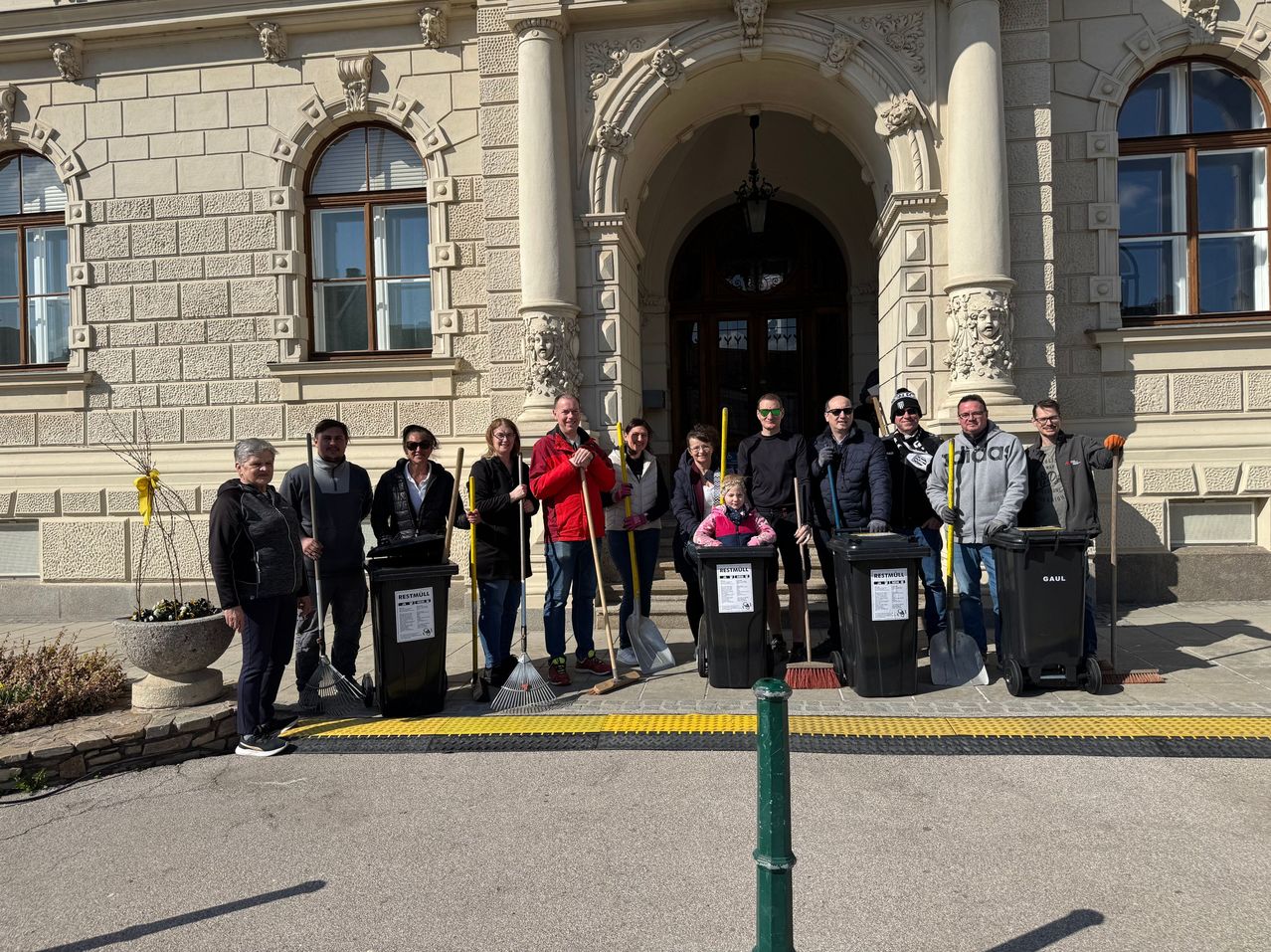 Eine Gruppe von Freiwilligen posiert mit Reinigungswerkzeugen vor einem Gebäude mit klassischer Architektur. Ein Kind sitzt in einem Mülleimer, und alle tragen Winterkleidung.