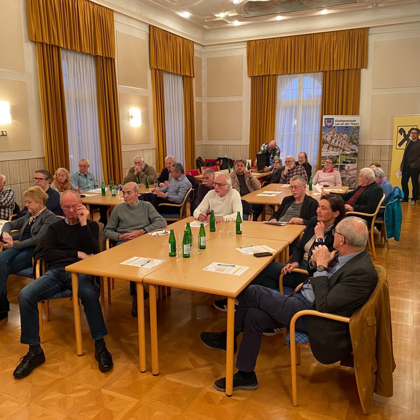 Eine Gruppe von Menschen sitzt an Tischen in einem Raum mit Holzböden. Flaschen und Papiere liegen auf den Tischen. Einige Personen stehen und eine Person geht. Banner hängen an der Wand.