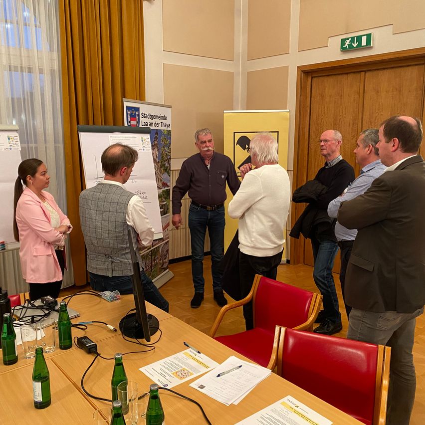 Eine Gruppe von Menschen steht um einen Tisch in einem Besprechungsraum. Eine Frau mit einem Pferdeschwanz schaut einen Mann an, der eine karierte Weste trägt. Ein Whiteboard und ein Poster sind in der Nähe.