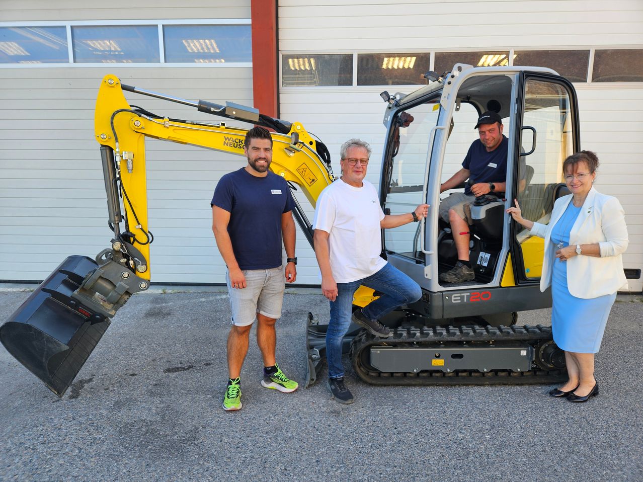Three men stand next to a yellow excavator, with one sitting inside. They are smiling and appear to be posing for a photo.