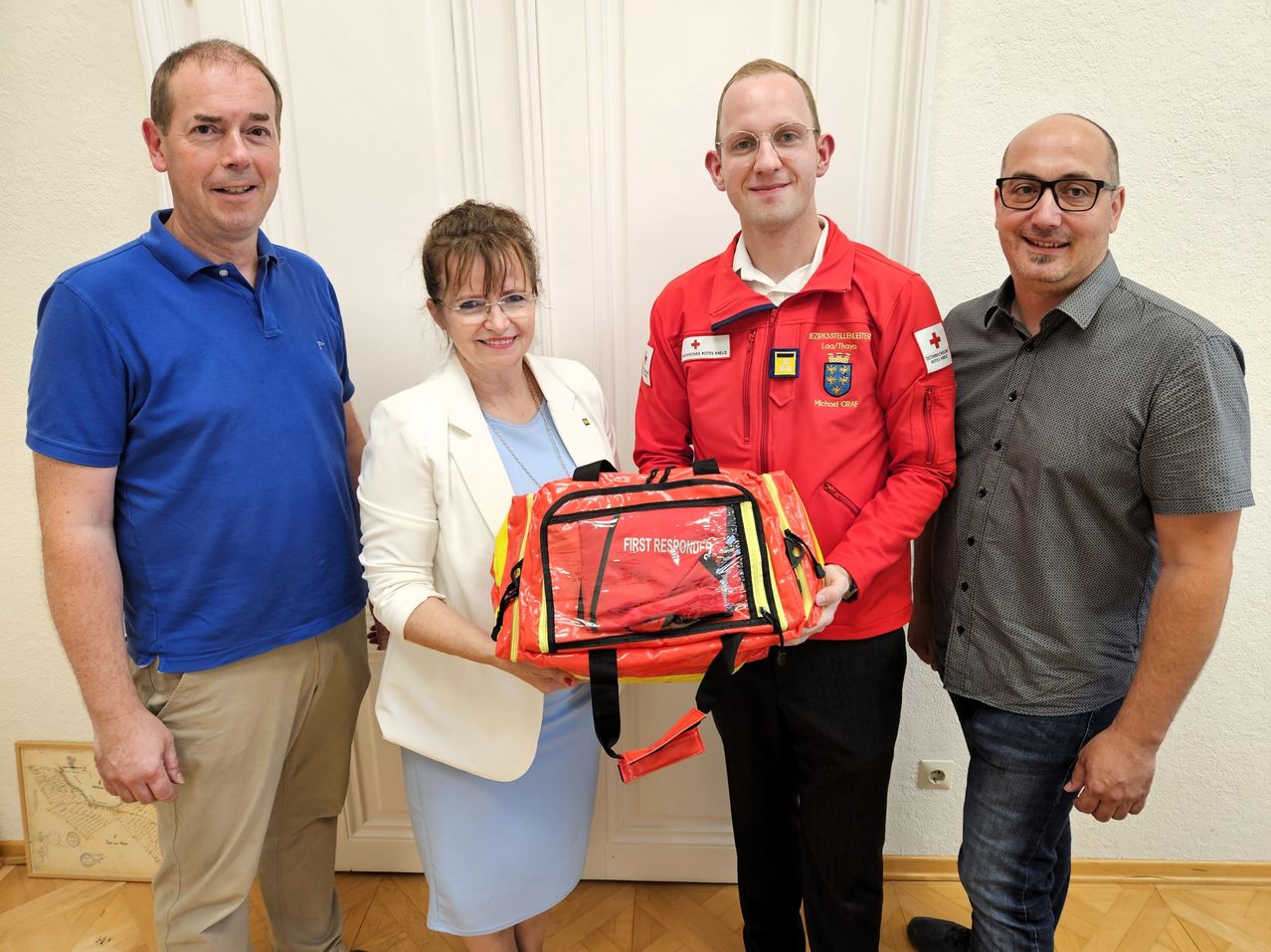 Four individuals stand together, smiling. A woman holds a red and yellow bag labeled 'First Responder'. The man next to her wears a red jacket with a logo and glasses. The man on the left wears a blue shirt and khaki pants. The man on the right wears a gray shirt and glasses.