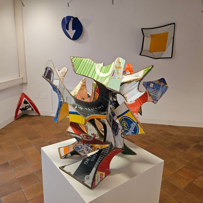 A modern art sculpture made from recycled cardboard and paper, displayed on a white pedestal in a gallery with tiled flooring.