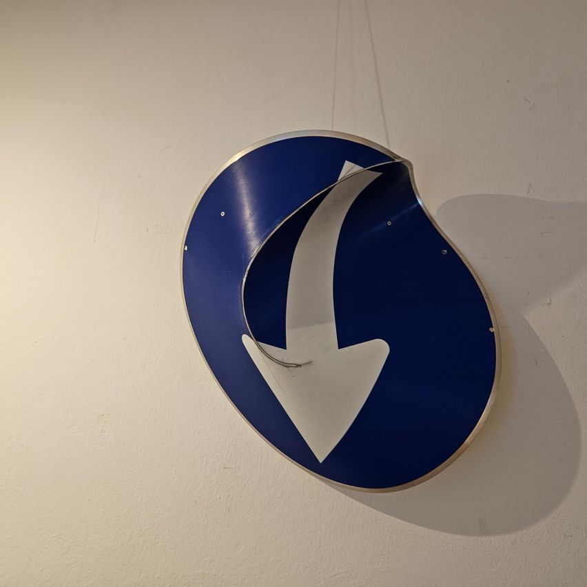A blue and white circular road sign with a white arrow pointing to the left is mounted on a white wall.