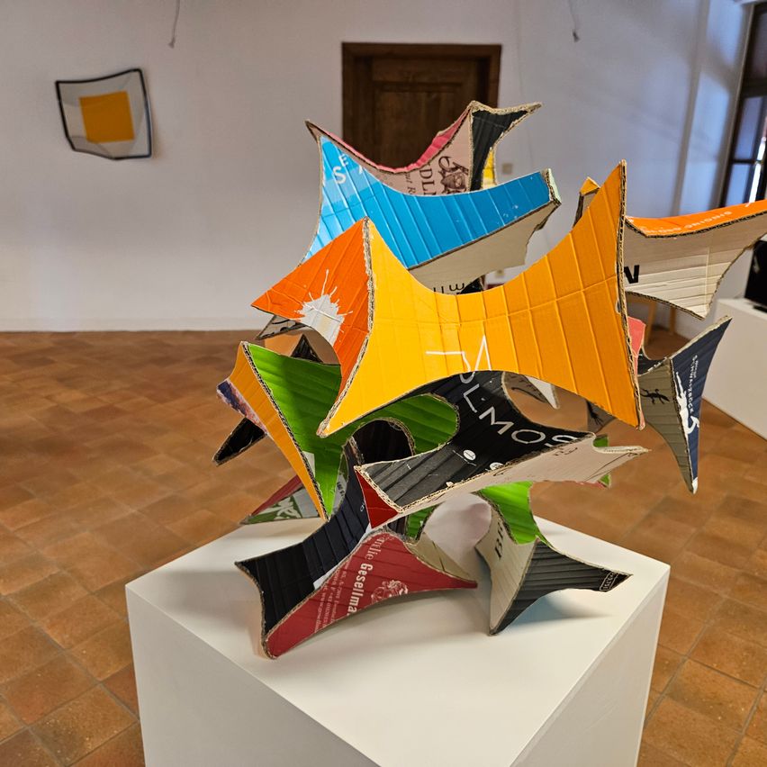 A vibrant sculpture made of colorful cardboard pieces, displayed on a white pedestal in an indoor setting with tiled flooring and a wooden door in the background.