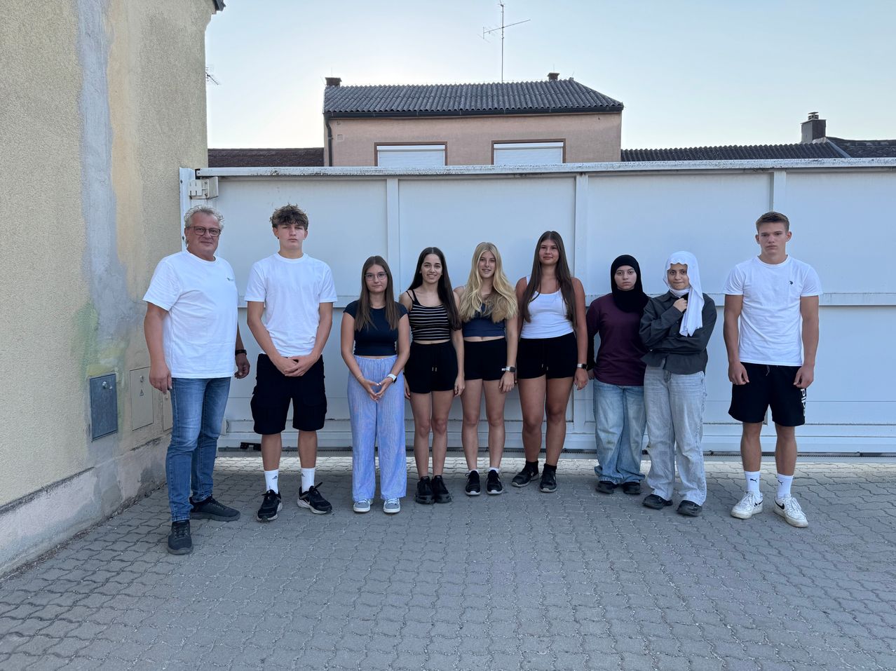 A group of young people and two adults stand in front of a white wall, posing for a photo. The setting is outdoors with a house in the background.